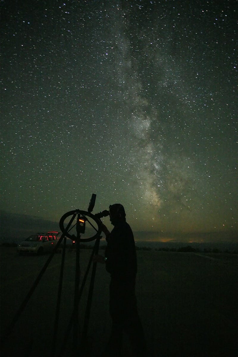 Ein nur als Silhouette erkennbarer Mann beobachtet in der Nacht mit einem Teleskop Sterne im Band der Milchstraße