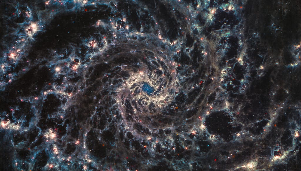 Eine Detailaufnahme der Galaxie Messier 74, in der Fotos bei unterschiedlichen Wellenlängen übereinandergelegt sind, zeigt die wirbelnden Spiralarme dieser Galaxie und in deren Zentrum einen bläulichen Sternhaufen.
